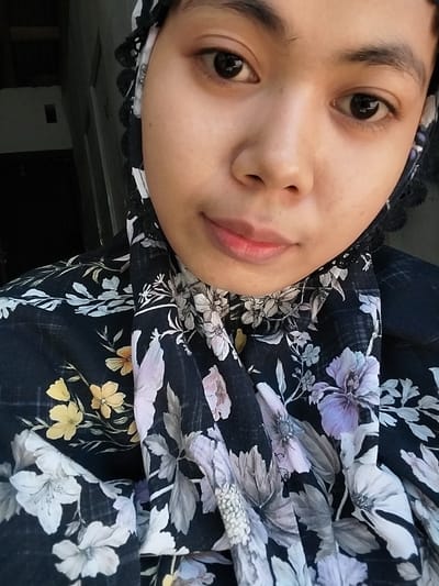 A close-up, selfie-style portrait of a young woman wearing a black floral-patterned hijab is shown. She is looking directly at the camera with a neutral expression. The background is dark and out of focus, with a hint of a doorway or opening to the left. The lighting suggests an indoor setting, possibly a home. The image is taken in Watampone, Indonesia, a city known for its vibrant culture and predominantly Muslim population, which aligns with the woman's attire.