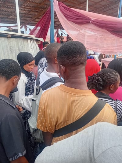 The image displays a crowded gathering occurring under a corrugated metal roof in Ogbogbo, Nigeria. Red and light pink sheer fabrics are draped and tied from the ceiling structure. On the far left, a corrugated metal partition stands next to a white vertical pole.
In the foreground, several individuals of African descent are standing. The leftmost person, viewed from behind, has short dark hair and wears a black collared shirt with white piping on the sleeves. Adjacent to them, a woman in a black hijab adorned with white dot embellishments and a white long-sleeved top over a dark patterned garment is looking downwards, revealing a nose piercing. To her right, another individual, seen from behind, wears a white long-sleeved shirt with a black and white patterned layer visible underneath. Further right, a person wears a mustard yellow short-sleeved t-shirt with vertical white stripes and has a black strap, possibly from a bag, across their shoulder. Partially visible behind this person is a woman with braided hair styled into a bun with a black tie, wearing a black and white striped top and an earring.
In the mid-ground and background, other attendees are seated on chairs, some dressed in traditional white robes and blue trousers. A table covered with a pink cloth is visible in the background. The individuals in the foreground are standing in close proximity, appearing to observe or wait.