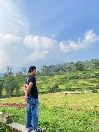 A person wearing a black T-shirt and blue jeans is standing in a rural area of Penanggungan, Indonesia. To the person's right is a green hillside area, and a rice paddy is visible in the background. Above, the sky is blue with white clouds. The person appears to be looking upwards. The image is oriented sideways.