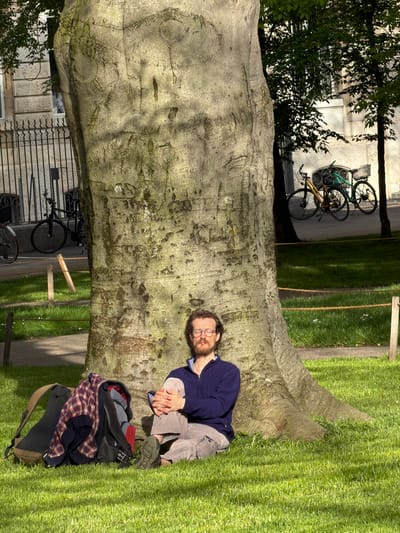 A man is depicted sitting on the ground, leaning against the wide, textured trunk of a large, light-barked tree. He has a beard, wears glasses, a navy blue long-sleeved top, and light brown or khaki pants, with his hands clasped in front of him. He appears to be looking towards the camera with a relaxed expression. Next to him, on the ground, rests a black and brown backpack, adorned with a folded red and blue plaid shirt and another dark garment.
The large tree trunk, which forms a prominent feature in the center of the image, has a somewhat mossy, weathered appearance. To the man's right, a vibrant green lawn extends into the background, where a simple wooden structure is visible. To his left, behind the tree, an urban setting is present, featuring a light-colored building with windows and dark iron railings. Several bicycles, including green, yellow, and black models, are parked upright in front of the building, suggesting a busy but calm street or pathway.
The scene is illuminated by bright, clear sunlight, indicating daytime and good weather conditions. The overall setting appears to be a park or public green space situated within an urban or suburban area, consistent with the location context of Basel, Switzerland. There is no visible text in the image.