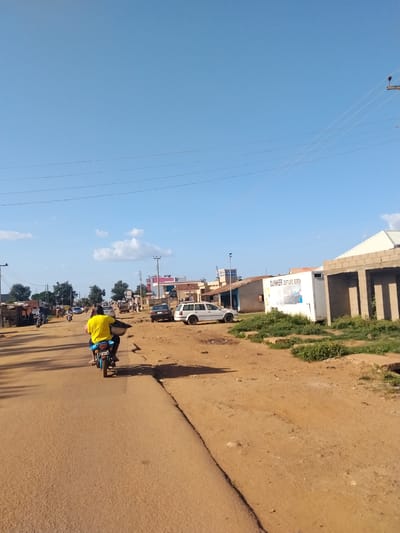 The image depicts a bustling street scene in Mangu Town, Nigeria, under a clear blue sky with a few scattered white clouds. The time of day appears to be morning or early afternoon, given the bright sunlight casting shadows. The road is unpaved and dusty, indicative of a rural or semi-urban setting.
The primary subject in the foreground is a person riding a motorcycle, wearing a bright yellow t-shirt and blue shorts. They are carrying a large, round object in front of them. The motorcycle has a license plate, though the numbers are not clearly legible. Several other vehicles, including cars and motorcycles, are visible on the road and parked along the sides, suggesting moderate traffic. Buildings line both sides of the street, some appearing to be shops or homes with corrugated iron roofs. One building prominently displays a white rectangular sign with text that reads "DUINKER GESTUURD BOREN" in black lettering.
The scene conveys a sense of everyday activity and commerce. People are seen walking and standing around, some gathered in small groups. The overall atmosphere is lively and dynamic, characteristic of a market day or a busy thoroughfare. The natural environment is represented by sparse vegetation and trees, adding to the impression of a community situated in a somewhat arid or developing region. The presence of power lines stretched across the sky indicates access to electricity.