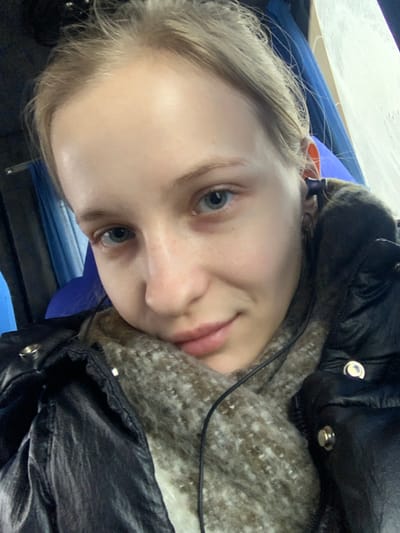 A close-up portrait depicts a fair-skinned female, approximately young adult, looking directly forward with a subtle smile. Her light blonde hair is pulled back, revealing her forehead. She has light blue eyes with subtle dark circles present beneath them, and her lips are slightly parted. She is dressed in a dark, possibly black, puffer-style jacket, beneath which a thick, textured, mottled beige and brown scarf is visible, wrapped around her neck. A black wired earbud is present in her right ear, with the wire extending downwards. A silver-toned snap fastener is visible on the right side of her jacket. The background is out of focus, showing indistinct blue upholstery or fabric to the left and a light-colored, possibly frosted surface or partition to the right, consistent with an interior setting, potentially a vehicle. The scene is located in Khodosivka, Ukraine.