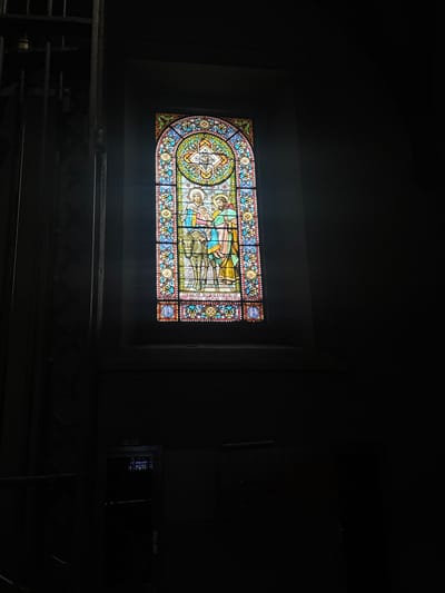 The image shows a stained-glass window depicting a religious scene, likely the Nativity, within a dimly lit interior space. The window is arched at the top and features figures of Mary, Joseph, and a baby, accompanied by a donkey. The top of the window has a cruciform design with angelic figures. Intricate patterns and borders, composed of various colors like blue, red, yellow, and green, frame the central figures. Light filters through the stained glass, illuminating parts of the artwork. To the left of the window, dark, vertical bars suggest a grate or railing, contributing to the somber atmosphere. In the lower portion of the image, a dark cabinet or door with a handle is visible, further shrouded in shadow. The overall environment appears to be a church or a similar sacred building, with a focus on the detail and artistry of the stained glass. The location is the Church of Sant Esteve in Collbató, Spain, as indicated by the text on the stained glass which includes "SANCT ESTEVE" and a coat of arms.