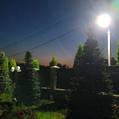 This image depicts an outdoor scene at twilight in Tohatin, Moldova. The foreground is dominated by a patch of grass with several rose bushes, some with visible red and pink blooms. Several evergreen trees, likely fir or cypress, are prominent throughout the scene. A brick fence is visible behind some of the trees, topped with a stone cap. Two spherical lights are mounted on posts along the fence line. In the mid-ground, a tall, white lamppost with a bright, diffused light source is positioned to the right of the frame, illuminating the surrounding area and creating lens flare. Power lines are visible stretching across the dark blue and gradient orange twilight sky. The background consists of a dense line of trees, suggesting a wooded area or dense foliage. The overall atmosphere is tranquil, with artificial light sources contrasting with the fading natural light. No people are visible in the image.