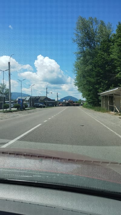 The image captures a sunlit view from inside a vehicle looking down a paved road in Teresva, Ukraine. The sky is bright blue with scattered white cumulus clouds.
On the left side of the road, utility poles with streetlights line the shoulder, beyond which are several buildings, including one with a visible Pepsi sign. A dark-colored SUV is parked near these structures. In the distant background on the left, mountains rise under the clear sky.
The road itself is asphalt, featuring a solid white line on the left edge and a dashed white line indicating the center or a lane divider. On the right side of the road, a dense line of tall green trees runs parallel to the pavement. Further down on the right, there is a simple, rustic structure, possibly a bus stop or shelter, made of wood with a white roof. The immediate foreground shows the dashboard and windshield of the vehicle from which the photograph was taken. The overall scene suggests a rural or semi-urban environment on a clear, sunny day. No people are visible.