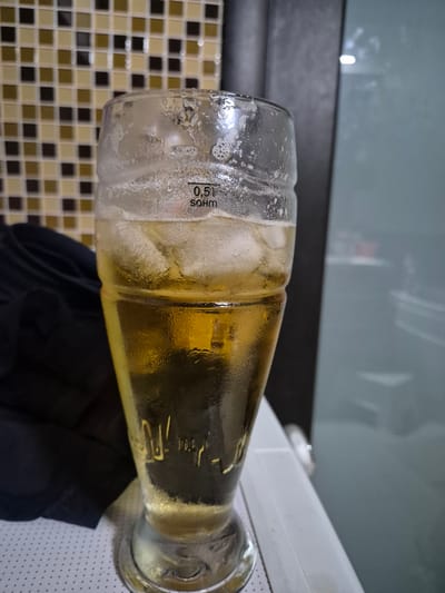 A glass bottle, filled with a golden-colored liquid and ice, lies on its side. The bottle is partially filled, with condensation visible on the exterior. A measurement marking on the bottle reads "0,5L". The background shows a tiled wall with a checkered pattern of brown and beige squares. Above the tiled wall, a grey surface is visible, possibly a countertop or shelf, with reflections of light. A dark piece of fabric is draped underneath the bottle. The scene appears to be indoors, possibly in a kitchen or bar area, given the tiled backsplash and the presence of a beverage. There is no indication of the time of day, weather, or specific emotions being conveyed. The location context of Nizhnevartovsk, Russia, is provided but not visually evident in the image itself.