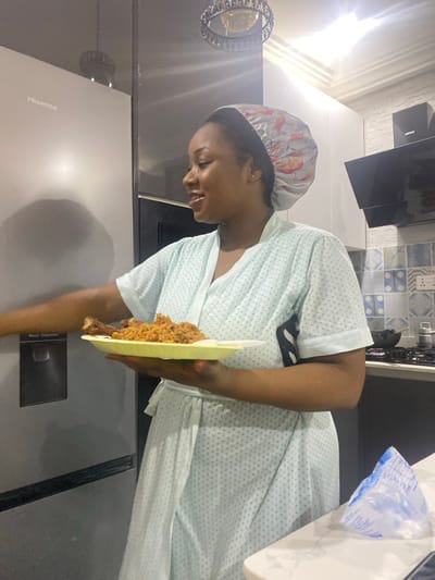 The image, rotated 90 degrees clockwise, presents a woman in what appears to be a kitchen or dining area. She is focused on a disposable foam plate in her hand, which holds a serving of a reddish-brown rice dish, likely jollof, accompanied by a piece of chicken. The woman is wearing a light-colored, short-sleeved robe or house dress with a subtle dotted pattern, and a patterned shower cap or sleeping bonnet on her head. Her expression is calm, with her gaze directed downwards.
The background reveals typical kitchen elements. To the left, a large, silver-grey Hisense refrigerator is visible, with the brand name "Hisense" and the label "Water Dispenser" distinctly legible on its surface. Above the refrigerator, a decorative, circular light fixture with reflective or crystalline elements adds a touch of interior design. In the upper background, patterned blue and white tiles adorn a wall, suggesting a backsplash or accent. Further to the right, a part of a dark kitchen cabinet or appliance is seen. A white plastic bag with faint, illegible text is also partially visible on the far right. The scene is brightly lit by indoor lighting.