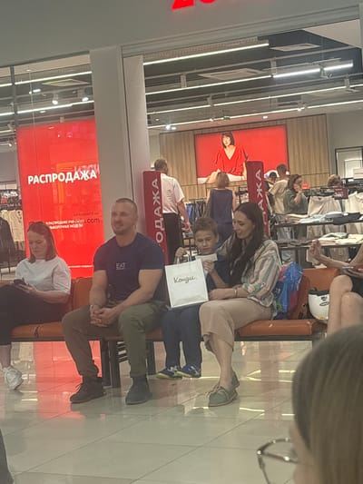 The image depicts a scene inside a retail store, likely a clothing store, with a prominent red display in the background featuring the word "РАСПРОДАЖА" (SALE) in large white letters. Several people are seated on an orange bench. In the foreground, a man with a shaved head and a dark blue t-shirt with an "EA7" logo is looking to his right, his hands clasped in front of him. Next to him, a woman with long dark hair, wearing a striped shirt and beige pants, is holding a white shopping bag with a logo that appears to say "Концепт." She is looking down and to her left, seemingly interacting with a young boy seated between her and the man. The boy is wearing a blue t-shirt and is also looking at the shopping bag. To the left of the man, a woman with red hair is looking at her phone. In the background, a screen displays an image of a woman in a red dress. Racks of clothing are visible, and other shoppers are scattered throughout the store. The floor is tiled and reflective. The overall impression is that of a busy shopping environment.