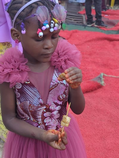 A young girl is depicted in a medium, eye-level shot, turned slightly to her left, with her head tilted towards the left, set against a vibrant reddish-pink background. Her skin tone is dark brown, and her hair is intricately braided, adorned with colorful beads and butterfly-shaped hair clips, some of which are translucent and iridescent. She is wearing a light pink headband. Her earlobes have small flower-shaped earrings, one purple and one pink.
She is dressed in a light pink dress with a ruffled, net-like material around the shoulders. The bodice features a burgundy fabric with a white floral and abstract pattern. Her hands are visible; the left hand is holding something that appears to be food, possibly a piece of fried yam or tofu, on a wooden skewer. Both hands show hints of an orange or red tint on the fingertips, possibly from henna or food coloring. She has a faint tattoo or mark on her right forearm.
In the background, on the far left, a person's legs and feet are visible, clad in dark pants and shoes. A dark wooden or metal railing is also partially visible behind them. The reddish-pink background appears to be a textured fabric, possibly a carpet or blanket, draped over a surface. Above the girl's right hand, a light-colored, possibly white, string or rope with a green and white tassel-like object is hanging down from the top edge of the frame. The lighting suggests it is daytime. The image captures a moment of focus or contemplation as the girl looks down at what she is holding.