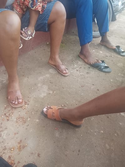 In Orodo, Nigeria, four individuals are present, with a focus on their feet and lower legs. One person is wearing blue trousers and appears barefoot with black slides. Another individual is wearing denim shorts, and only their legs are visible. A third person is partially visible, wearing brown slides and denim shorts. The fourth person is a child, wearing a colorful dress and small brown sandals. The scene is set outdoors on a concrete surface with scattered debris. A motorcycle is visible in the background to the right.