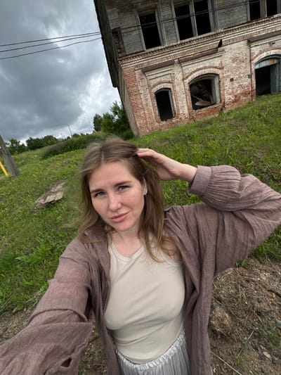 A young woman with shoulder-length blonde hair and blue eyes stands in front of a derelict brick building. She is wearing a beige tank top and a loose, brown, open-fronted cardigan with pleated sleeves. Her left hand is resting on the back of her head, and her right arm is bent at the elbow, out of view. The building behind her is made of red brick with large, empty window openings. The sky is overcast with dark clouds. The foreground is a grassy slope with some loose soil and debris. The location appears to be the outskirts of a village, possibly in rural Russia, given the style of the building and the surrounding vegetation.