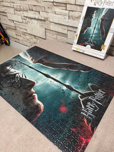 The image shows a completed Harry Potter puzzle on a textured, tan surface. The puzzle depicts a scene from "Harry Potter and the Deathly Hallows – Part 2" with Harry Potter and Lord Voldemort facing each other, wands extended. The puzzle is partially in focus, with the right side and top edge of the puzzle box visible in the upper right quadrant. The puzzle box is white with text and imagery related to the movie. In the background, a wall with a stacked stone tile pattern is visible. To the left of the puzzle, a black and neon yellow backpack is leaning against the wall. The scene is set on a flat surface, possibly a table or floor, which has a slight texture. The lighting suggests an indoor setting, possibly a room in Ko Kaeo, Thailand, given the context provided. There are no people actively engaged in the scene.