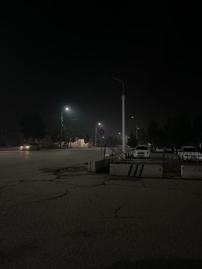 The image displays a nighttime street scene in Karmana, Uzbekistan. The overall environment is dark with low visibility, suggesting an atmospheric haze or fog.

In the immediate foreground, dark asphalt pavement covers the bottom third of the frame, exhibiting numerous cracks and several small, dark puddles. Three rectangular concrete barriers are positioned on the right side; one barrier, closer to the viewer, features three distinct black vertical stripes. Behind these barriers, a partially obscured metal gate or fence-like structure is visible.

The midground features an asphalt road that appears to split or form an intersection. On the left, a light-colored sedan is in motion, its headlights illuminated, casting a glow on the road ahead. On the right side of the road, multiple white sedans are parked parallel to the curb. A tall, white pole with a functional streetlight extends upwards on the mid-right, emitting bright light. Further down the road, closer to the center, another illuminated streetlight is visible. A circular regulatory sign, red with a blue cross (indicating "No Stopping" or "No Parking"), is affixed to a pole near the center-right.

The background consists of dark, silhouetted trees lining the sides of the road. Faint outlines of low-rise buildings are discernible through the darkness and atmospheric conditions behind the trees and parked vehicles. No people are visible in the scene.