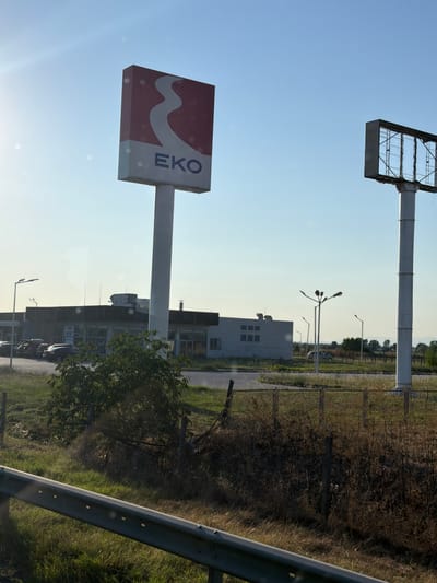 The image depicts an outdoor scene under a clear blue sky, likely during the day. In the foreground, a metal guardrail runs horizontally across the bottom of the frame, with a grassy area to its right and a road with some vegetation beyond it.
The main subjects are two large signs on tall poles. The sign on the left features the logo of "EKO," a stylized white wavy line against a red background, with the letters "EKO" in blue below it. The sign on the right is a large, empty billboard frame.
In the middle ground, there is a low-rise building with a dark-colored roof. A sign on this building reads "FOR RENT." Several cars are parked in front of the building. There are also streetlights visible, one of which has a unique, multi-armed design. A small, bushy tree is situated between the EKO sign and the building.
The background consists of a flat landscape with more trees and vegetation, suggesting a somewhat rural or suburban setting. The overall impression is of a commercial area. Based on the context provided, this scene is located in Radinovo, Bulgaria.