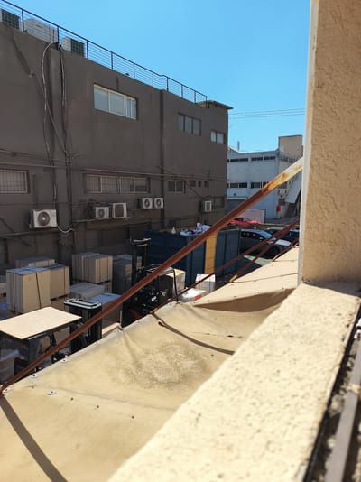 The image captures an outdoor industrial area in Azor, Israel, under a clear blue sky. In the foreground, a textured, light-colored wall and a weathered metal railing dominate the right side, suggesting a vantage point from a balcony or upper floor. The view looks down onto an area filled with stacked boxes, some wrapped in plastic, and pallets. A black forklift, with "TOYOTA" clearly visible on its mast, is positioned near the boxes. Further back, a large, dark blue industrial bin is visible, filled with debris. Beyond this, the facade of a dark gray building with multiple windows and air conditioning units is prominent. The building's roof is equipped with a black metal railing. In the distance, a lighter-colored building with many windows can be seen, and a red car and a dark-colored car are parked on a paved surface. The scene suggests a commercial or warehousing district.