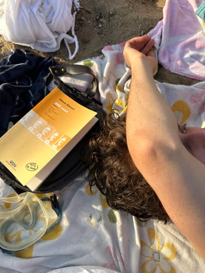 The image captures a close-up, overhead view of a person relaxing outdoors, likely on a beach. The main subject is partially visible, showing a person's right arm and the back of their head with dark, curly hair. The arm is tanned from the sun and appears to be resting on or near a towel. The person's head is turned away from the camera, and they seem to be lying down, facing away from the sunlight.

Around the person are various items indicative of a leisurely outdoor activity. The person is lying on a light-colored towel with yellow floral or leaf patterns, which is spread out over sand. Another pink and white patterned towel is visible to the left. To the left of the person's arm, on the sand, is a crumpled white piece of fabric, possibly a garment or towel.

Beneath the person's head and arm, a dark bag, possibly a shoulder bag or a small backpack, is placed on the patterned towel. Resting on top of this bag is an open book with a distinct orange-yellow cover. The book's title and author are clearly visible in Turkish: "Nâzım Hikmet" is the author, and "835 satır" (meaning "835 lines") is the title. Below the title, smaller text includes "1929'dan", "Bugünden", and "835 Satır I. Kitap". The cover also features a series of small black and white photographic portraits, likely of the author. A circular logo with "YKY" and "20. baskı" (20th edition) is visible, along with the publisher's name, "Yapı Kredi Yayınları."

To the right of the book and bag, a pair of light-colored swimming goggles or a snorkel mask with a dark strap is resting on the patterned towel. A dark blue and white striped fabric, possibly a piece of clothing like a t-shirt or swimwear, is partially visible beneath the bag.

The scene is brightly lit by natural sunlight, casting distinct shadows, suggesting it is daytime, possibly late morning or afternoon. The sandy ground confirms a beach or similar coastal environment. The overall impression is one of relaxation and leisure on a sunny day.
