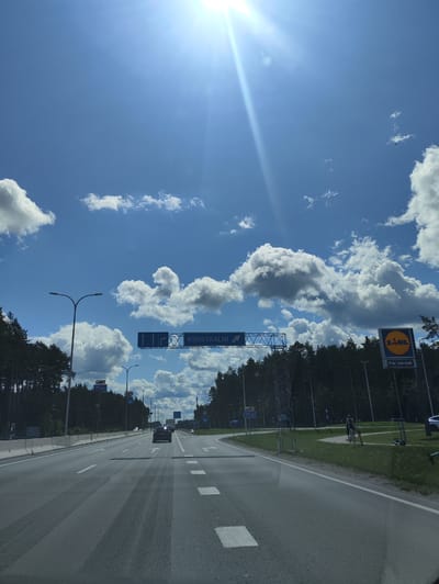 The image shows a view from a vehicle traveling on a highway towards Krustkalni, Latvia. The sky is bright blue with scattered white clouds, and the sun is shining intensely. On the right side of the road, a Lidl store sign is visible. The highway is paved with asphalt and has white lane markings. Trees line the sides of the highway. There are a few cars on the road, and a person is riding a bicycle on the grassy area beside the road.