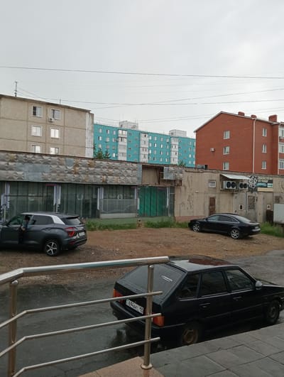 The image depicts an outdoor scene in what appears to be a residential or mixed-use area, likely in Kostanay, Kazakhstan, given the license plate. The weather is overcast, suggesting a cloudy or rainy day, as indicated by the wet pavement in the foreground and the general dull lighting.
In the foreground, a wet paved area with a metal railing suggests a walkway or entrance. A black, older model hatchback car is parked on the right, with its license plate visible: "KZ 694AKL 10". A sticker is visible on its rear windshield with text that reads "AUTOSLEGION TATYANAKAMYSLOVA.RU".
Behind this car, on a slightly elevated, dirt-covered area, a dark gray SUV is parked with its driver's door open and a person visible inside. This SUV also has a license plate. Further back, a long, low building with a metallic, textured facade and large windows runs horizontally across the mid-ground. Various air conditioning units and some metal grating are visible on its roof.
In the background, two apartment buildings are prominent. To the left, a lighter-colored, older apartment building with several windows and air conditioning units is visible. To the right of that, a taller, distinctively turquoise-colored apartment building with many windows and balconies stands out. To the far right, a reddish-brown brick building with multiple stories and windows is visible, suggesting a mix of architectural styles and building ages in the area. The overall impression is of a somewhat utilitarian urban environment under grey skies.