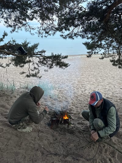 The image depicts two individuals on a sandy beach, likely in Ozerki (Krasnodolinskaya), Russia, under the shade of pine trees. The scene is set near the Baltic Sea, evidenced by the calm water and distant horizon. A small campfire is burning in the foreground, fueled by dry branches. One person, wearing a hooded jacket and camouflage pants, is tending to the fire. The other individual, wearing a vest over a long-sleeved shirt and a red, white, and blue baseball cap, is also engaged with the campfire, holding a stick. A black thermos or canister is visible hanging from a tree branch in the upper left portion of the frame. The sand is lightly textured with footprints and debris from the surrounding vegetation. The sky is overcast, suggesting diffused natural light.