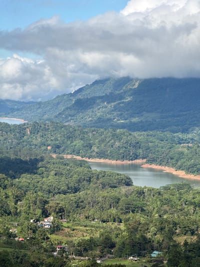 Lush green mountains covered in dense forests stretch across the landscape, partially veiled by cloud formations. A body of water, likely a lake or river, meanders between the mountains. Patches of bare earth are visible along the water's edge. Lower down, structures, possibly houses or buildings, are interspersed within the trees, hinting at human habitation. The scene is captured from a high vantage point on a partly cloudy day.