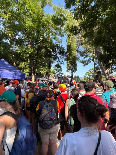 The image depicts a crowd of people gathered outdoors, likely at the Circuit de Barcelona-Catalunya in Montmeló, Spain, given the presence of racing-themed attire. The foreground shows several individuals from behind, wearing casual clothing such as t-shirts, shorts, and hats. Many are sporting colorful backpacks, and some hats feature prominent logos, including "aramco" and "PETRONAS." The crowd is dense, suggesting an event or gathering.
In the background, more people are visible, along with tents and trees. The environment appears to be a sunny day with a clear blue sky visible through the foliage. The overall scene conveys a sense of a public event or festival, with a focus on fans attending an outdoor venue.