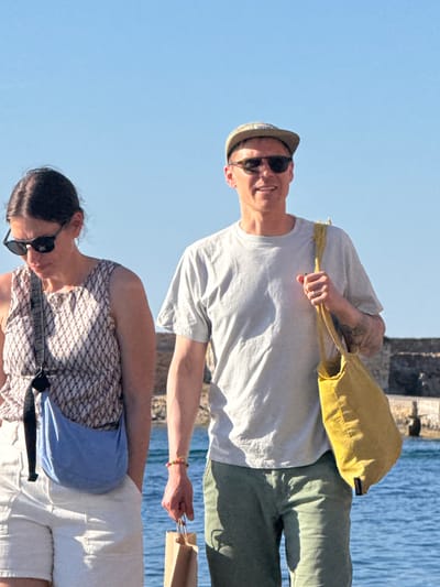 Two individuals, a woman and a man, are walking outdoors near the water. The woman on the left is wearing white shorts, a patterned sleeveless top, and sunglasses. She has a blue bag slung across her body. The man on the right is wearing green shorts, a light grey t-shirt, sunglasses, and a cap. He is carrying a yellow tote bag over his shoulder and a brown paper bag in his hand. They are positioned against a backdrop of a clear blue sky and the blue water of the sea. In the background, a stone structure is visible. The scene appears to be captured in Chania, Greece, possibly along the Venetian Harbor or a similar coastal area.