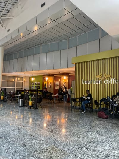 The image captures an indoor scene featuring a "Bourbon Coffee" shop situated within a larger, modern public building. The coffee shop is prominently marked by a yellow and wood-slat facade with the words "bourbon coffee" written vertically in white, accompanied by a stylized leaf logo.

Inside the coffee shop, several people are visible, mostly seated at tables, suggesting they are patrons. One person, dressed in a dark top and blue jeans, stands at a high table near the coffee shop's entrance. Backpacks and a maroon duffel bag are visible near some seating areas. The interior is warmly lit by pendant lights and recessed fixtures, and decorated with potted greenery.

Adjacent to the coffee shop is a spacious open area with a light-colored, possibly tiled or stone, floor that shows reflections of interior lighting. The ceiling of this larger space is composed of a grid of white and gray panels with integrated lighting, indicating a contemporary architectural design.

The activity taking place is typical of a coffee shop: people are relaxing, possibly socializing or working. The setting appears to be a public transit hub, mall, or large commercial building in Kigali, Rwanda, as suggested by the "Bourbon Coffee" brand, which is known in the region. The scene is illuminated by artificial lights, creating a bright indoor environment.