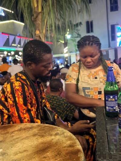 A man and a woman are seated at an outdoor table in a dimly lit area. The man, on the left, wears a vibrant orange and black patterned shirt and black beaded necklaces. He has a beard and is looking down, holding a phone in his right hand. The woman, on the right, wears a light-colored patterned shirt with geometric designs. Her hair is braided. She is also looking down, seemingly engaged with something on the table. A green bottle of Star beer is positioned between them on the table, which appears to be made of a woven material. In the background, palm trees and lights suggest a tropical or semi-tropical urban setting at night. Other people are visible in the background, indicating a public space such as a restaurant or bar. The overall atmosphere is casual and social. The location is identified as Oba, Nigeria.