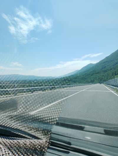 The image depicts a view from a vehicle traveling on a highway. The highway has two lanes in the direction of travel, marked with white lines. A metal guardrail runs along the right side of the road. On the left side of the highway, there is a lower guardrail and beyond that, a valley with scattered buildings and trees. In the background, a lush green mountain range dominates the landscape under a clear blue sky with some wispy white clouds. The foreground shows the dashboard and windshield of the vehicle, with a textured mat on the dashboard. A motorcycle is visible on the highway in the distance, in the right lane. The location is identified as being near Leonessa, Italy.