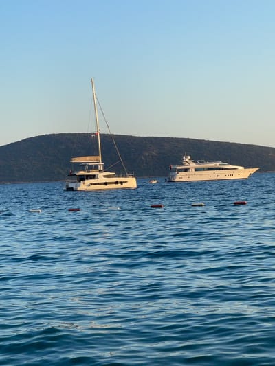 Two yachts are moored in the clear blue waters off the coast of Bodrum, Turkey, under a cloudless sky. The larger vessel on the right is a sleek, white motor yacht with multiple decks and windows. To its left, a white catamaran sailboat with two hulls is anchored, its mast reaching high into the sky. The background features a lush green, tree-covered hillside. In the foreground, several buoys mark the water's surface. No people are visible on the yachts or in the immediate vicinity. The scene is peaceful and serene, characteristic of a tranquil day at sea.
