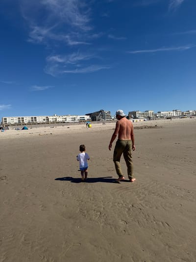 A father and son are walking on the sandy beach in Berck, France. The son, wearing a white t-shirt and blue shorts, is walking ahead of his father. The father, shirtless and wearing khaki pants and a baseball cap, is walking behind his son. In the background, buildings line the coastline, and a few beach umbrellas and tents are visible. The sky is a clear blue with wispy clouds. The sun is casting shadows on the sand.
