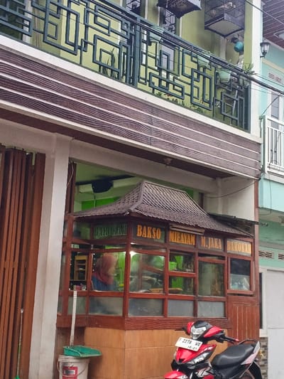 The image depicts an urban street scene in Pinnwe, Myanmar (Burma), featuring a two-story building. The upper floor is light green with a balcony equipped with a dark green metal railing displaying a complex geometric pattern. Several birdcages and potted plants are positioned on this balcony. The ground floor includes a brown wooden facade, a white structural column, and a prominent street-level wooden food stall. This stall features a brown pagoda-style roof, glass panels, and exterior signage that reads "BAROKAH," "BAKSO," "MIE AYAM," "M. JAKA," and "BAROKAH." Inside the stall, an individual wearing a reddish-brown head covering and blue clothing is visible. A red and black scooter is parked directly in front of the stall, displaying license plate F 2278 WO. To the immediate left of the stall, a white bucket with red markings, a green dustpan, and a white pole are situated on the pavement. Further left, a brown wooden accordion-style gate is partially visible. Overhead electrical wires traverse the upper portion of the frame, and a section of a light blue building facade is visible to the right.