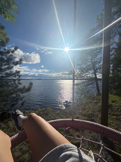The image shows a first-person perspective of someone sitting in a pink woven swing, looking out at a calm lake on a sunny day. The sun is high in the sky, casting bright rays and creating a glare on the water. Trees and foliage frame the scene, with a thin wooden post visible on the right side. The person's legs, wearing shorts and sandals with socks, are prominent in the foreground. The environment appears to be a natural, wooded area near a body of water, likely in the Unorganized Thunder Bay District, Canada, given the suggestion of a northern landscape with coniferous trees. The overall atmosphere is relaxed and serene, suggesting a moment of leisure in nature.