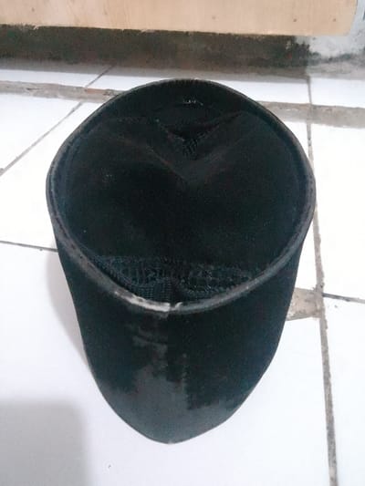 A black, kufi-style hat is positioned on a tiled floor. The hat is cylindrical with a flat top, and the material appears to be fabric. The top of the hat has some stitching details creating a pattern. The hat is the sole object in the foreground, and its placement suggests it has been set down. The background shows a floor composed of white square tiles, with grout lines visible. Above the tiled floor, there's a wooden surface, possibly a shelf or part of a structure, which is lighter in color. The lighting suggests an indoor setting, possibly with natural light filtering in. The scene is still and lacks any human presence or apparent activity, focusing solely on the presentation of the hat. There is no visible text in the image. The location context is Cisarua, Indonesia.