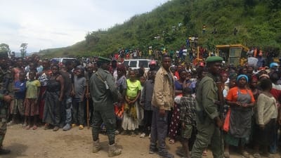 The image shows a large crowd of people, mostly women and children, standing on a dirt road in Sake, Democratic Republic of Congo. There are two military personnel standing amongst the crowd, one of them holding a rifle. In the background, there are several vehicles and a large yellow construction machine. The scene is chaotic, with people pushing and shoving to get a better look. The image shows a sense of tension and unrest. The people in the crowd appear to be looking towards the vehicles and construction equipment, suggesting that they are waiting for something to happen. There is a hill with lush vegetation in the background. The location of the image is in a rural area of Sake, on a dirt road with a clear sky. The temperature is likely hot. The image appears to have been taken during the daytime. The mood is tense and expectant, with the crowd looking on with curiosity and perhaps fear.