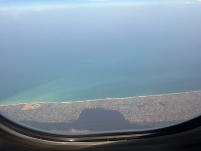 The image shows a view from an airplane window overlooking a coastal area. The ocean fills the upper portion of the frame, transitioning to a shoreline with a dense urban environment along the coast. The city is likely Cotonou, Benin, based on geographical cues and the nature of the urban layout visible.

Specifically, the shoreline exhibits a narrow sandy beach followed by densely packed buildings and infrastructure stretching inland. There is a prominent dark area in the lower central portion of the window view, likely caused by a shadow or reflection within the window itself. The water color shifts from a deeper blue further out to a lighter turquoise near the shore, indicating shallow water. The sky is bright with some hazy conditions near the horizon.