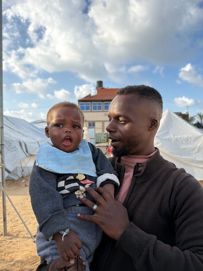 An adult male, identified as Black, holds an infant, also identified as Black, in an outdoor setting in a-Zawayda, Palestinian Territory. The man, with short dark hair and a beard, wears a dark brown zippered jacket over a pink collared shirt and a silver ring on his left ring finger; he looks slightly down and to his right. The infant, wearing a grey long-sleeved top with a multi-colored design, grey sweatpants, and a light blue terry cloth bib, has an open mouth and faces directly forward. The man supports the infant with both hands. In the background, under a partly cloudy blue sky, multiple white canvas tents or temporary shelters are visible on a sandy ground, along with a two-story building featuring a red tile roof and multiple windows.