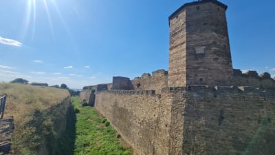 This media file captures a sun-drenched view of the Bilhorod-Dnistrovskyi fortress in Ukraine. The scene is dominated by the ancient stone walls and a prominent tower of the fortress, bathed in bright sunlight that creates lens flare effects in the upper left corner. The sky is a clear, vibrant blue with a few scattered white clouds.

The fortress walls are constructed from rough-hewn stones and appear weathered, suggesting great age. Horizontal bands of red brick are visible interspersed with the stone in the main tower, adding a distinctive pattern. Crenellations top the walls, and remnants of battlements are visible.

In the foreground, a dry moat is visible on the left, filled with tall, dry grass. Beyond the main wall, a verdant green area with shorter grass suggests a different part of the fortress grounds or its surroundings. The perspective looks out over the fortress walls and down into the moat area. In the distance, beyond the fortress, a body of water can be seen, hinting at a coastal or riverine location. Silhouetted trees and some low buildings are also visible in the background, indicating the presence of a settlement near the fortress.

The bright sunlight and clear sky suggest it is midday on a fair-weather day. The lack of people or any ongoing activity points to a peaceful, historical site. The overall mood is one of historical grandeur and a sense of timelessness, enhanced by the bright, clear conditions. There is no visible text in the image.