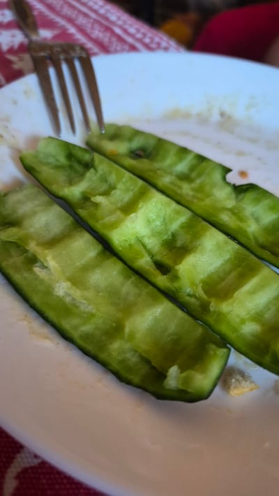 The media file displays a close-up, overhead view of sliced cucumbers on a white plate, set against a red and white patterned tablecloth. A fork with three tines is visible in the lower left corner, partially resting on the plate and the tablecloth. The cucumbers are a vibrant green, with visible seeds and flesh, suggesting they have been recently cut. There are a few crumbs scattered on the plate, hinting at a meal or snack being consumed. The lighting appears to be natural and from an overhead source, casting subtle shadows. The overall scene conveys a casual, domestic setting, possibly a kitchen or dining area in Krauja, Latvia. No individuals are clearly visible, but the presence of food and cutlery suggests a meal in progress. The image captures a simple, everyday moment focused on the food.