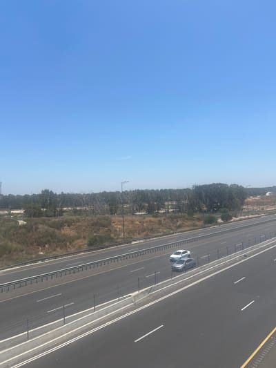 The image shows a highway with two lanes of traffic in each direction, running through a semi-arid landscape near Hadera, Israel. The sky is clear and blue. On the highway, there are two cars: a white sedan in the left lane and a gray sedan in the right lane. The highway is bordered by dry vegetation and trees, suggesting a warm, possibly summer season. A metal guardrail separates the lanes of traffic, and a concrete barrier with a wire fence runs along the side of the road. Streetlights are visible in the distance. The overall scene is one of a typical highway environment in a relatively undeveloped area.
