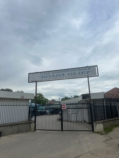 This is a photograph of the entrance to "PIATA AGROINDUSTRIALĂ ROADA GLIEI" in Strășeni, Moldova. A large white sign with blue text, mounted on a pole, displays the name of the market and its address: Str. Serghei Lazo 1-a. Below the name, contact telephone numbers are listed. The sign is tilted, with the text running vertically.
The entrance itself is secured by a black metal gate, which is partially open, allowing a view into the market. Inside the gate, several vehicles, possibly cars and vans, are visible parked. The setting appears to be an outdoor market or a commercial area.
To the right of the gate, a concrete pathway leads away from the entrance. A red and white circular sign with a horizontal bar, indicating "no entry" or a prohibition, is affixed to the gate.
The sky above is overcast with significant cloud cover, suggesting a cloudy or potentially rainy day. The overall lighting is diffused, indicating it might be daytime, but the exact time is not discernible. The photograph captures a moment of everyday activity at a commercial hub in Strășeni.