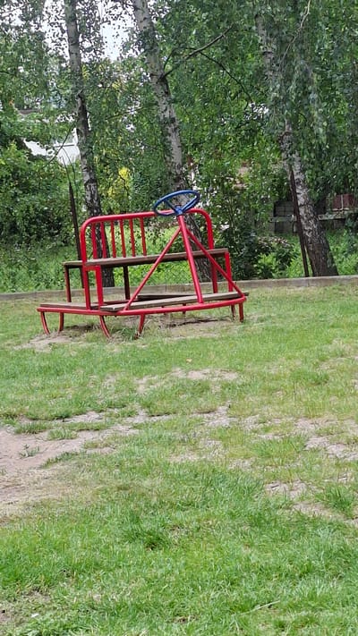 A red metal playground ride with wooden seats and a blue steering wheel is situated on a grassy area. The ride appears to be a type of miniature vehicle or carriage. It is surrounded by trees and vegetation. The ground is covered in patchy green grass with some bare earth. In the background, there is a fence and a building. This image was taken in the city of Grójec, Poland.