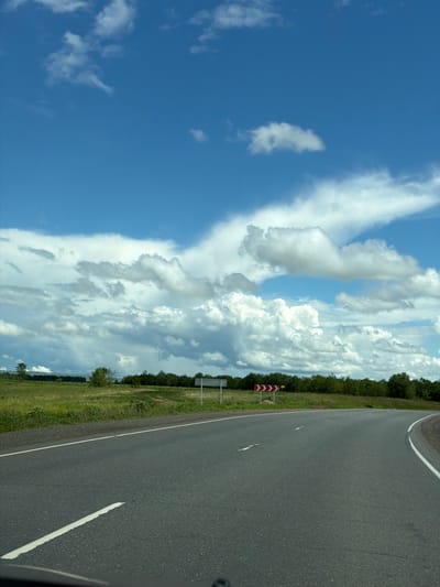 The image depicts a highway curve with a bright blue sky filled with dramatic white cumulus clouds overhead. The road itself is asphalt with white lane markings, curving to the right. To the left of the road, a grassy field is visible, bordered by a line of trees in the distance. There are a couple of road signs along the side of the road, one with red and white chevron markings indicating a curve. No people or vehicles are present in the image. The scene appears to be set in a rural or semi-rural area.