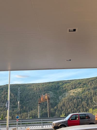 The image shows a building, likely a commercial or industrial structure, situated in Kalotina, Bulgaria, adjacent to a highway with a car parked on the shoulder. The building is a light neutral color with a long, uniform facade. There are two small window or vent openings visible. The highway is separated from a hillside covered with dense green trees by a guardrail. There are power lines and related infrastructure visible on the hillside. The sky is light blue with some scattered clouds.