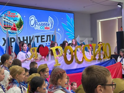 In Pron'kino, Russia, a group of young people stand behind a large banner displaying the text "Орлята России" (Orylyata Rossii - Eagles of Russia) and "Хранители" (Khraniteli - Guardians). They are holding large, gold-colored letters that spell out "РОССИИ" (Rossii - of Russia). Several individuals in the foreground have Russian flag colors painted on their cheeks. The background features a large screen displaying graphics related to the event. The atmosphere appears to be festive and patriotic.