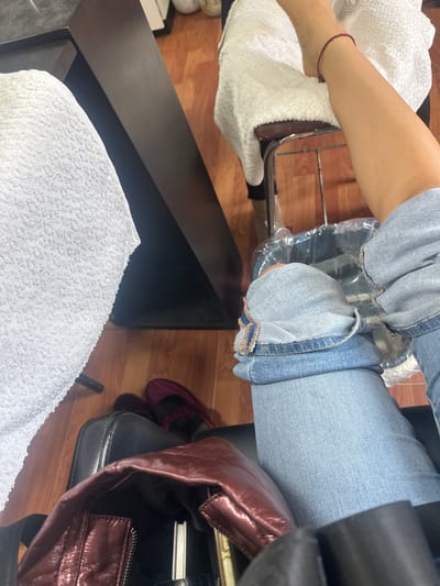 The image depicts a person receiving a pedicure in an indoor setting. The person is wearing light blue denim jeans and has one foot wrapped in a white towel. The other leg is resting on a chair with a plastic liner. A black table with another white towel on it is visible near the foot receiving the pedicure. A handbag and shoes are seen near the chair. The wooden flooring is visible in the background. This could be a salon, likely in the city of San Pedro Cholula, Mexico, assuming that information is accurate and relevant to the scene.