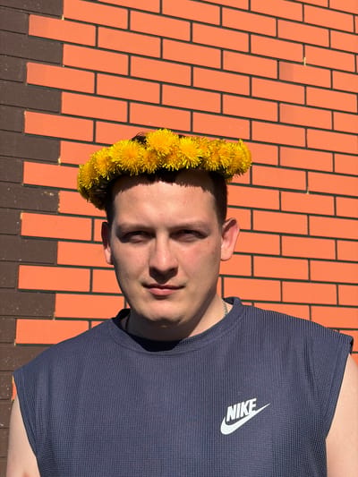 A person wearing a yellow dandelion crown is positioned on a brick surface. The person has short, dark hair and is wearing a navy blue Nike t-shirt. The background consists of light orange and dark brown bricks. The location is Кемуль, Russia.