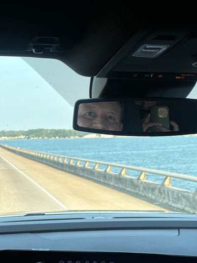 The image captures a first-person perspective from inside a vehicle looking forward through the windshield. The vehicle's interior features a black headliner and an overhead console with illuminated indicators, including "ON" and "PASS AIR BAG ON." A rearview mirror is centrally positioned. Reflected in the left side of the rearview mirror is the upper face of a male individual, appearing to be the driver. Reflected in the right side of the rearview mirror is a hand holding a smartphone with a multi-lens camera, pointed towards the mirror, indicating the photograph was taken by a passenger.
Through the windshield, a concrete bridge extends into the distance over a large body of calm, blue water. The bridge features at least one visible lane delineated by a solid white line, and a concrete barrier with a light-colored, repeating horizontal railing system along its right side. In the far background, across the water, a distant shoreline is visible with green vegetation and scattered structures. The sky above is clear and light blue.
The specific location of this bridge and expansive body of water cannot be definitively identified as being within the city of Montgomery, United States, based on the visual information provided.