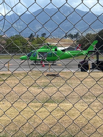 A bright lime-green helicopter, registered TI-BXX, is stationary on an asphalt helipad. Its main rotor blades are not in motion. The forward left cabin door of the helicopter is open. Two orange traffic cones are positioned on the asphalt near the helicopter's front landing skid. A dark-colored utility vehicle is partially visible, parked to the right and slightly behind the helicopter's tail. The helipad surface features yellow and white markings, including the numbers "24-10" faintly visible. The scene is viewed through a chain-link fence, which occupies the lower and central foreground. Beyond the fence, the ground consists of dry, yellowish-brown grass. The background features a line of green trees, some exhibiting pink blossoms, leading to a range of densely vegetated mountains under a partly cloudy sky. This location is identified as the Tobías Bolaños International Airport (MRPV) in Pavas, San José, Costa Rica.