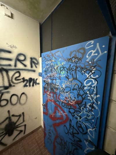 The image shows a low-angle view of an interior space with graffiti on the walls and ceiling. The ceiling is painted blue and covered in dense graffiti in black, white, and red spray paint. Some discernible words and phrases include "BITE!!", "LE MARIITE", "KLYANA AMB", "BISON BB", "Nique mo", and "Tanmede mei grosse mot". Below the ceiling, the walls are painted a dingy white, also covered in graffiti, including "RR" and "2PAC" in large black letters, as well as numbers like "600" and a spider drawing. The environment suggests a neglected, possibly abandoned, urban setting. The location, Amarante, Portugal, cannot be confirmed from the visual data alone.